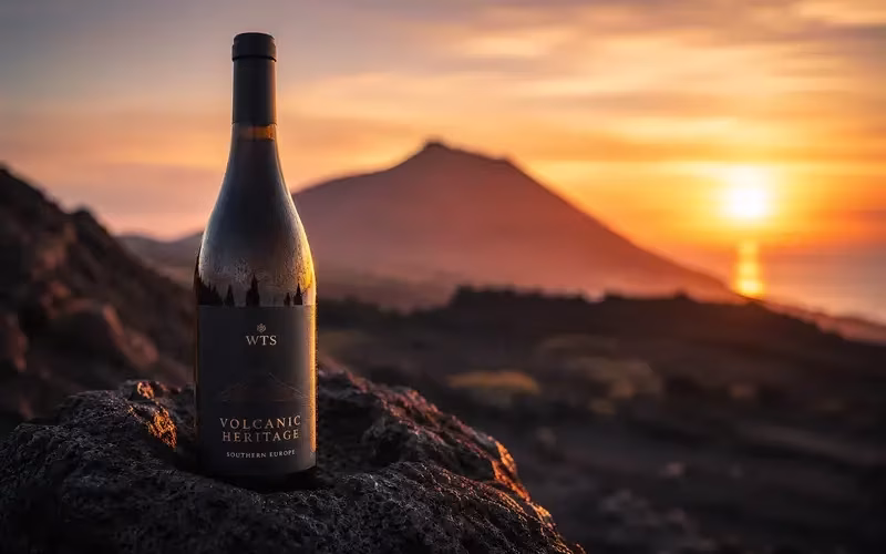 Premium wine bottle displayed on a volcanic lava rock at sunset, with a softly blurred volcano in the background, symbolising the unique heritage of Southern Europe's volcanic vineyards. C'est la version que je recommande pour ton article : elle est descriptive, élégante et reprend n